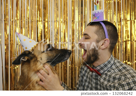 A playful man and his German Shepherd dog in party hats face each other during a fun birthday celebration. Pet birthday party concept 132068242