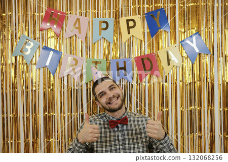 Cheerful man in party hat and red bow tie smiles with thumbs up in front of Happy Birthday banner. Front view portrait. Birthday party concept 132068256