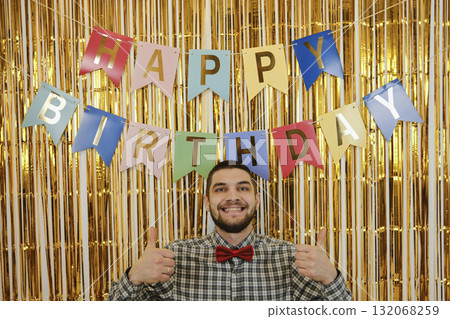 Smiling man in front of colorful Happy Birthday banner, wearing red bow tie, showing thumbs up 132068259