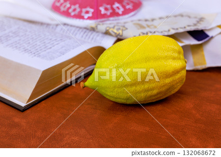Etrog citrus fruit on open prayer book religious text with traditional Jewish holiday symbols including kippah tallit for Sukkot festival celebration. Etrog citrus fruit on open prayer book religious text with traditional Jewish holiday symbols including kippah tallit for Sukkot festival celebration. 132068672