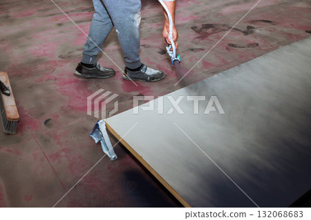 Worker is applying paint to wooden board using spray gun while standing on dusty floor Worker is applying paint to wooden board using spray gun while standing on dusty floor 132068683