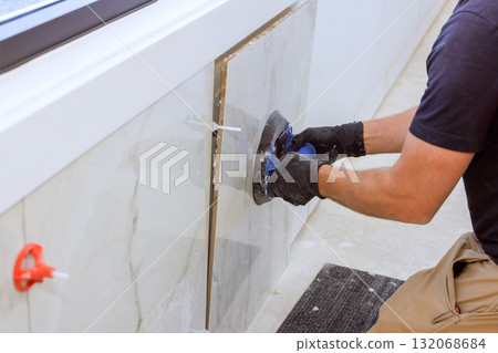 Construction worker carefully applies tile to wall with vacuum suction cup tool in spacious room. Construction worker carefully applies tile to wall with vacuum suction cup tool in spacious room. 132068684