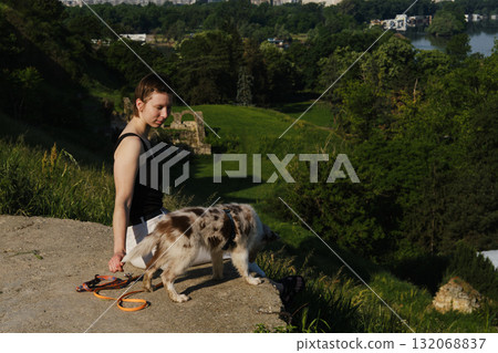 Woman sits on cliff edge with red merle Border Collie puppy on leash, enjoying view of green valley and city 132068837