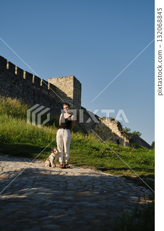 Young woman stands with red merle Border Collie puppy on leash by old Kalemegdan fortress wall, training and bonding outdoors. Travel with dog concept Young woman stands with red merle Border Collie puppy on leash by old Kalemegdan fortress wall, training and bonding outdoors. Travel with dog concept 132068845