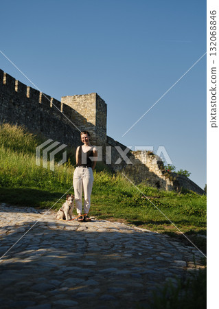 Young woman stands with red merle Border Collie puppy on leash by old Kalemegdan fortress wall, training and bonding outdoors. Travel with dog concept 132068846