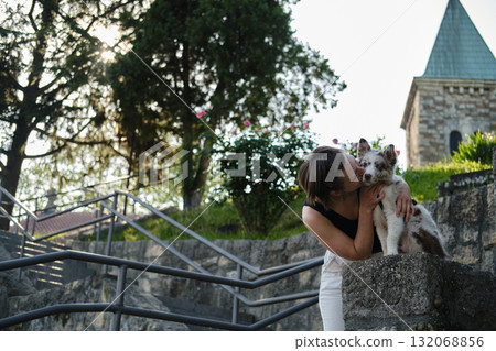 Woman kisses red merle Border Collie puppy sitting on stone wall in park, tender and affectionate outdoor moment. Travel with dog concept Woman kisses red merle Border Collie puppy sitting on stone wall in park, tender and affectionate outdoor moment. Travel with dog concept 132068856