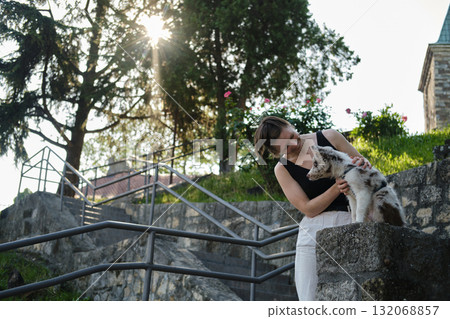 Young woman pets red merle Border Collie puppy on stone wall with sun rays shining through trees, outdoor bonding. Travel with dog concept Young woman pets red merle Border Collie puppy on stone wall with sun rays shining through trees, outdoor bonding. Travel with dog concept 132068857