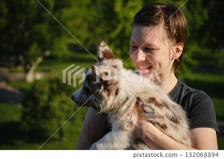Smiling young woman holds red merle Border Collie puppy in arms outdoors, enjoying warm sunshine and green park background 132068894