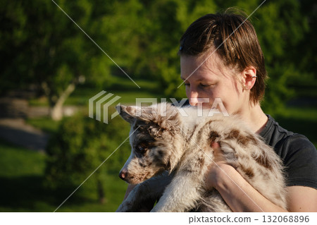 Smiling young woman holds red merle Border Collie puppy in arms outdoors, enjoying warm sunshine and green park background 132068896