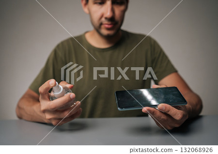 Selective focus portrait of man spraying sanitizer onto mobile phone screen, highlighting importance of hygiene and disinfection in everyday life on gray table surface. Concept of phone maintenance 132068926