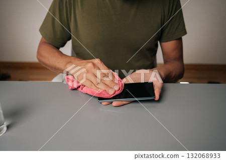 Close-up cropped shot of unrecognizable man wiping smartphone display with microfiber cloth, ensuring hygiene and maintaining clean device at home office workspace. Close-up cropped shot of unrecognizable man wiping smartphone display with microfiber cloth, ensuring hygiene and maintaining clean device at home office workspace. 132068933