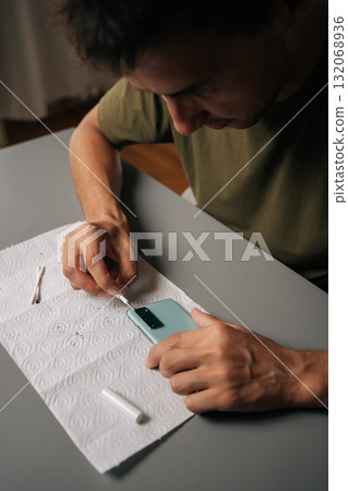 High-angle view of focused technician cleaning smartphone using specialized tools sitting at table, ensuring optimal quality and device performance. Concept of mobile phone maintenance and service. High-angle view of focused technician cleaning smartphone using specialized tools sitting at table, ensuring optimal quality and device performance. Concept of mobile phone maintenance and service. 132068936