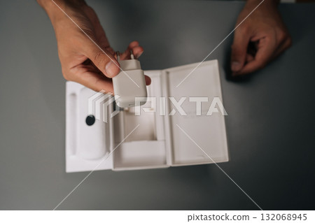Top view of young man unpacking holding white smartphone charger being unpacked from cardboard box, sitting on gray table. Concept of contemporary gadget unboxing. Top view of young man unpacking holding white smartphone charger being unpacked from cardboard box, sitting on gray table. Concept of contemporary gadget unboxing. 132068945