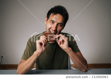 Portrait of happy Caucasian man sitting at table biting chocolate, savoring rich flavor of dark chocolate bar, blissful enjoyment as takes satisfying and delicious bite, on isolated background. 132068961