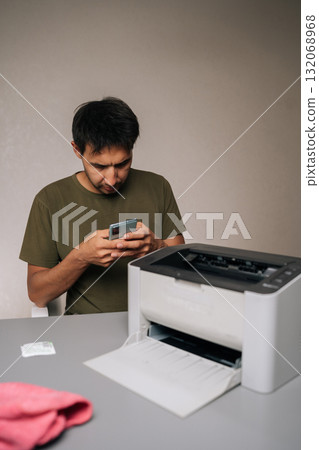 Focused man using smartphone to read instructions while repairing laser printer, with cleaning supplies and tools nearby on table, on isolated background. Concept of hardware, repairing, upgrade 132068968