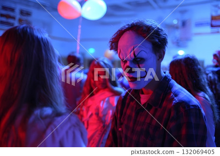 A spooky clown with face paint stands in a crowd on Halloween. A spooky clown with face paint stands in a crowd on Halloween. 132069405