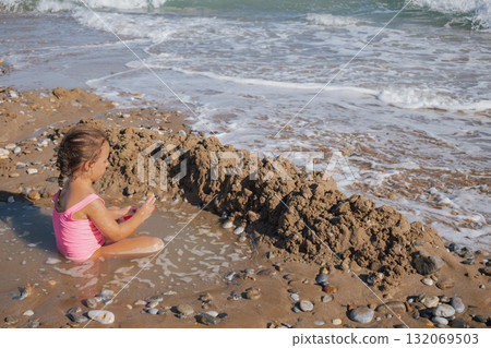 Happy Little Girl in Pink Swimsuit Building Sand Castle Moat on a Pebble Beach with Waves and Sea Foam in Summer Sunlight 132069503