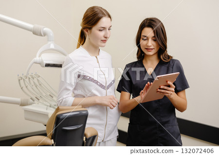 Two women dentist doctors are standing in a dental office and looking at a tablet 132072406