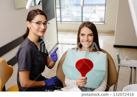 Woman is happy with results after oral hygiene and teeth whitening at dentist office. Patient is smiling and looking at the mirror 132072418