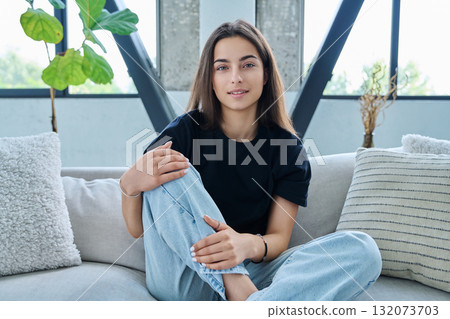 Beautiful young woman, sitting on sofa in living room at home, looking at camera Beautiful young woman, sitting on sofa in living room at home, looking at camera 132073703