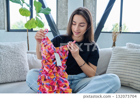Young woman unpacking cardboard box parcel with online order, looking at new clothes Young woman unpacking cardboard box parcel with online order, looking at new clothes 132073704