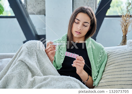 Sad upset teenage girl looking at thermometer holding blister pack of pills sitting at home Sad upset teenage girl looking at thermometer holding blister pack of pills sitting at home 132073705
