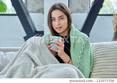 Beautiful young woman sitting under warm blanket with cup in her hands, on sofa Beautiful young woman sitting under warm blanket with cup in her hands, on sofa 132073706