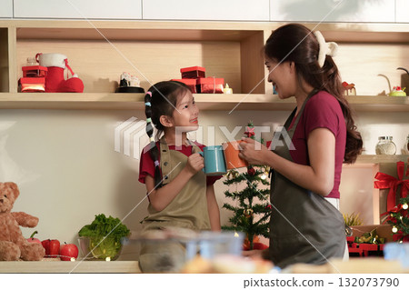 Holiday Cheer. Mother and daughter sharing joyful moment with mugs in festive kitchen. Holiday Cheer. Mother and daughter sharing joyful moment with mugs in festive kitchen. 132073790