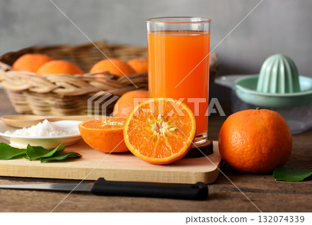 Half an orange is placed on a wooden chopping board. Placed on an old wooden table prepared for making orange juice, In the back there are oranges in the basket and the juicer container. Half an orange is placed on a wooden chopping board. Placed on an old wooden table prepared for making orange juice, In the back there are oranges in the basket and the juicer container. 132074339