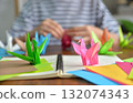 A close up shot of a bird folded from colorful origami paper with a blurred background of a woman folding the paper on wooden table top 132074343