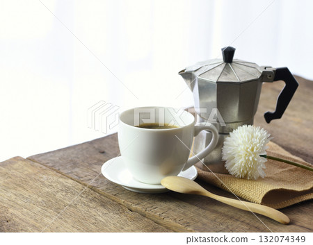 Black coffee in a white coffee cup Placed on an old wooden table by the window with a bright white curtain in the background, decorated with a moka pot, wooden spoon and sackcloth. Black coffee in a white coffee cup Placed on an old wooden table by the window with a bright white curtain in the background, decorated with a moka pot, wooden spoon and sackcloth. 132074349