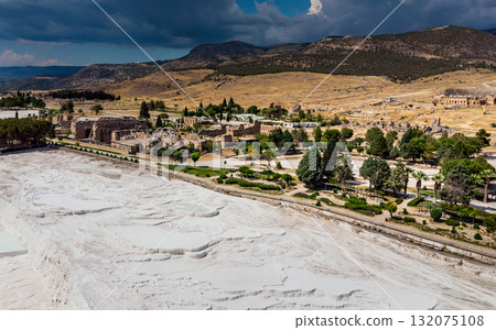 wide-angle landscape of Pamukkale's white travertine terraces and ancient Hierapolis ruins in Turkey under a dramatic sky. 132075108