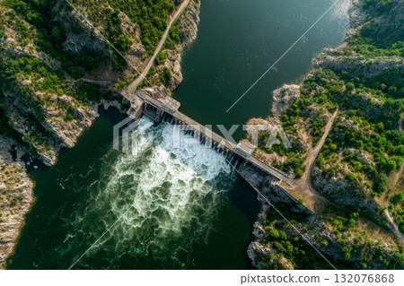 Hydroelectric dam with water flowing into a river between rocky green hills 132076868