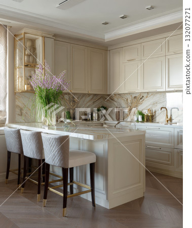 High-end, light-filled kitchen with classic cream cabinetry, striking gold-veined marble countertop and backsplash. The space features island with three velvet, quilted bar stools and brass hardware 132077271