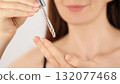 Close-up of an unrecognizable woman holding a pipette with facial serum. A drop of transparent oil or gel drips from the glass dropper. Skincare, cosmetic product, liquid collagen, beauty routine. 132077468