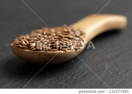 flax seeds poured into a wooden spoon on black slate , black surface with a wooden spoon filled with flax seeds 132077885