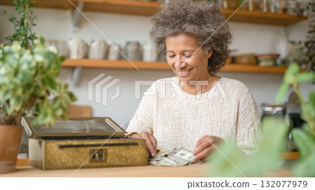 Happy older woman counting cash by a decorative box in a cozy, rustic setting. Represents personal finance, savings, small business, profit, retirement, money management, and success 132077979