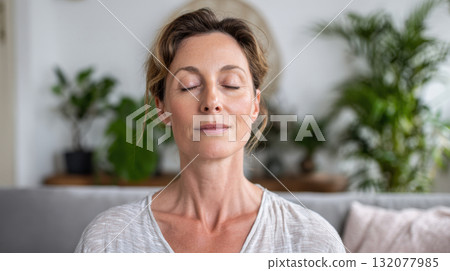 Peaceful middle-aged woman with closed eyes engaging in meditation, yoga, or mindful breathing indoors. Promoting mental health, relaxation, and a balanced lifestyle in a tranquil home environment 132077985