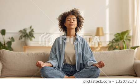 Horizontal shot of a peaceful young woman sitting cross-legged on a couch with her eyes closed and hands in mudra position, meditating. The bright, minimalist living room creates a calm atmosphere 132078178