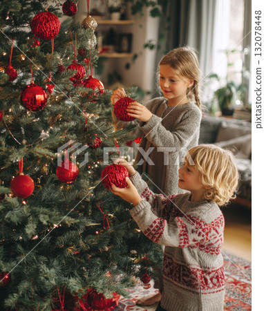 Two smiling children, a girl and a boy, are happily decorating a lush Christmas tree with vibrant, glittering red ornaments in a warm, inviting living room. Image beautifully captures joy, tradition 132078448