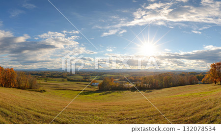 Stunning panoramic view of a rural valley in autumn, featuring golden fields, rolling hills, and forests ablaze with fall color under a brilliant sunburst and a wide, partly cloudy blue sky 132078534