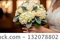 A hyper-realistic, high-detail close-up photograph of a bride's hands gently holding a gorgeous wedding bouquet filled with creamy white Hydrangeas, ivory Spray Roses, Dusty Mille 132078802