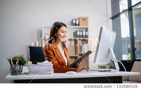 Young Asian woman, smiling and working at her desk in an office environment. She is wearing a business suit using a laptop. Young Asian woman, smiling and working at her desk in an office environment. She is wearing a business suit using a laptop. 132080326