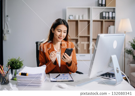 Young Asian woman, smiling and working at her desk in an office environment. She is wearing a business suit using a laptop. Young Asian woman, smiling and working at her desk in an office environment. She is wearing a business suit using a laptop. 132080341