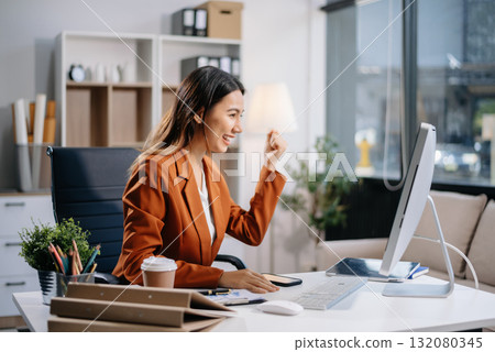 Young Asian woman, smiling and working at her desk in an office environment. She is wearing a business suit using a laptop. Young Asian woman, smiling and working at her desk in an office environment. She is wearing a business suit using a laptop. 132080345