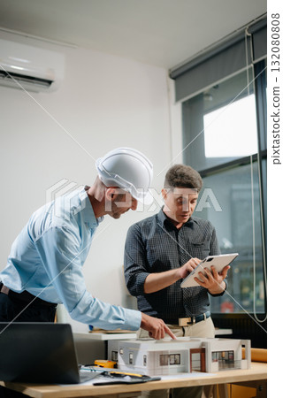 Professional collaboration in architecture. Two engineers work on construction plans, using tools like laptops, tablets, and scale models. Professional collaboration in architecture. Two engineers work on construction plans, using tools like laptops, tablets, and scale models. 132080808