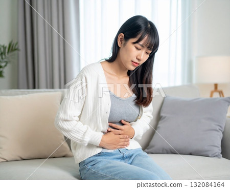 A Japanese woman holding her stomach at home and looking distressed A Japanese woman holding her stomach at home and looking distressed 132084164