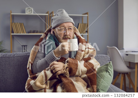 Senior man sitting wrapped in a blanket and trying to warm himself with a cup of hot tea. 132084257