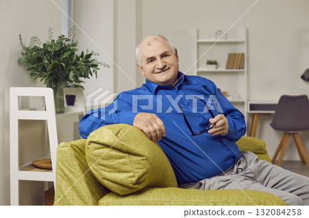 Portrait of an elderly man sitting on sofa in the living room at home and holding glasses in hands. 132084258