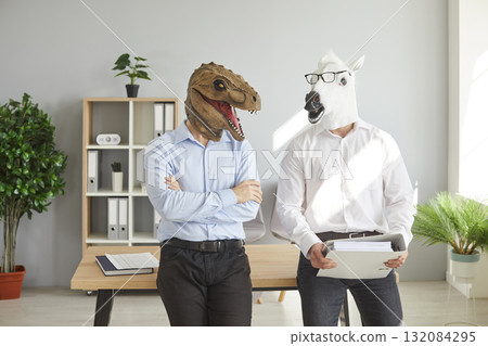 Two office workers in funny dinosaur and horse masks standing by desk and talking Two office workers in funny dinosaur and horse masks standing by desk and talking 132084295